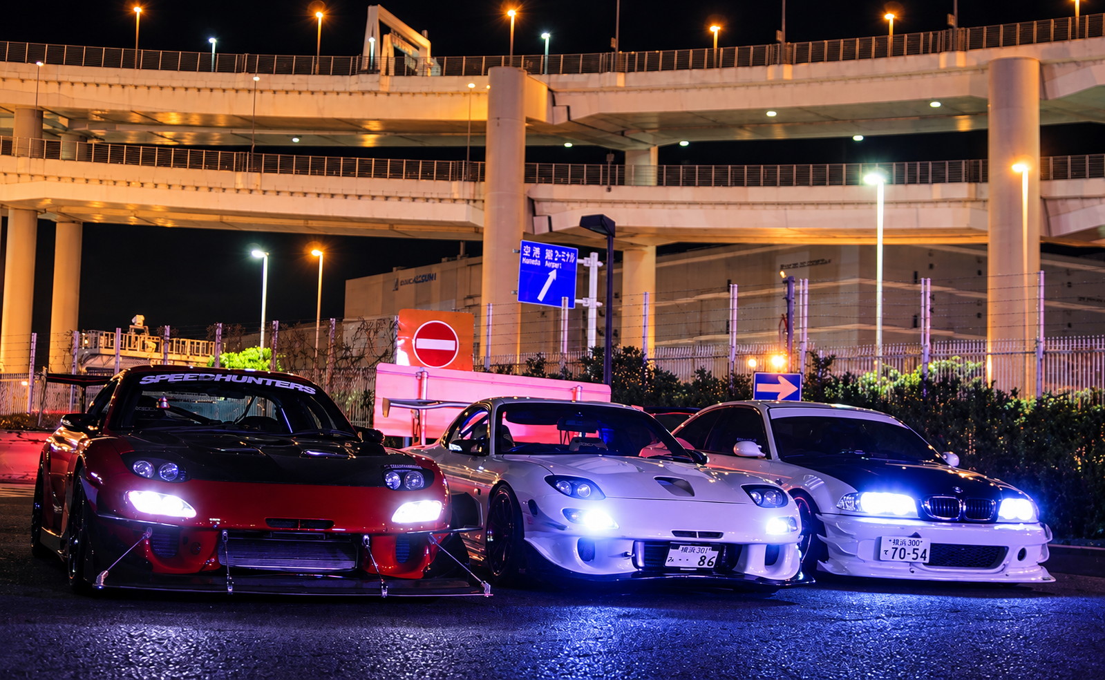 Tres deportivos japoneses reunidos de noche bajo una red de autopistas urbanas